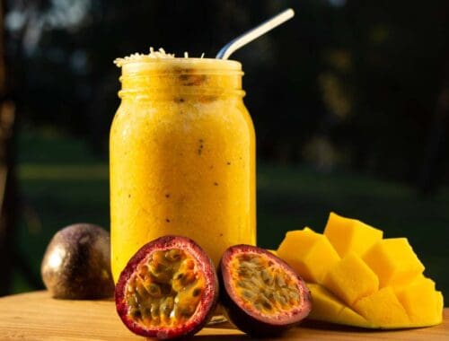 Fresh passion fruit and mango smoothie in a glass jar with a straw, displayed outdoors on a wooden surface.