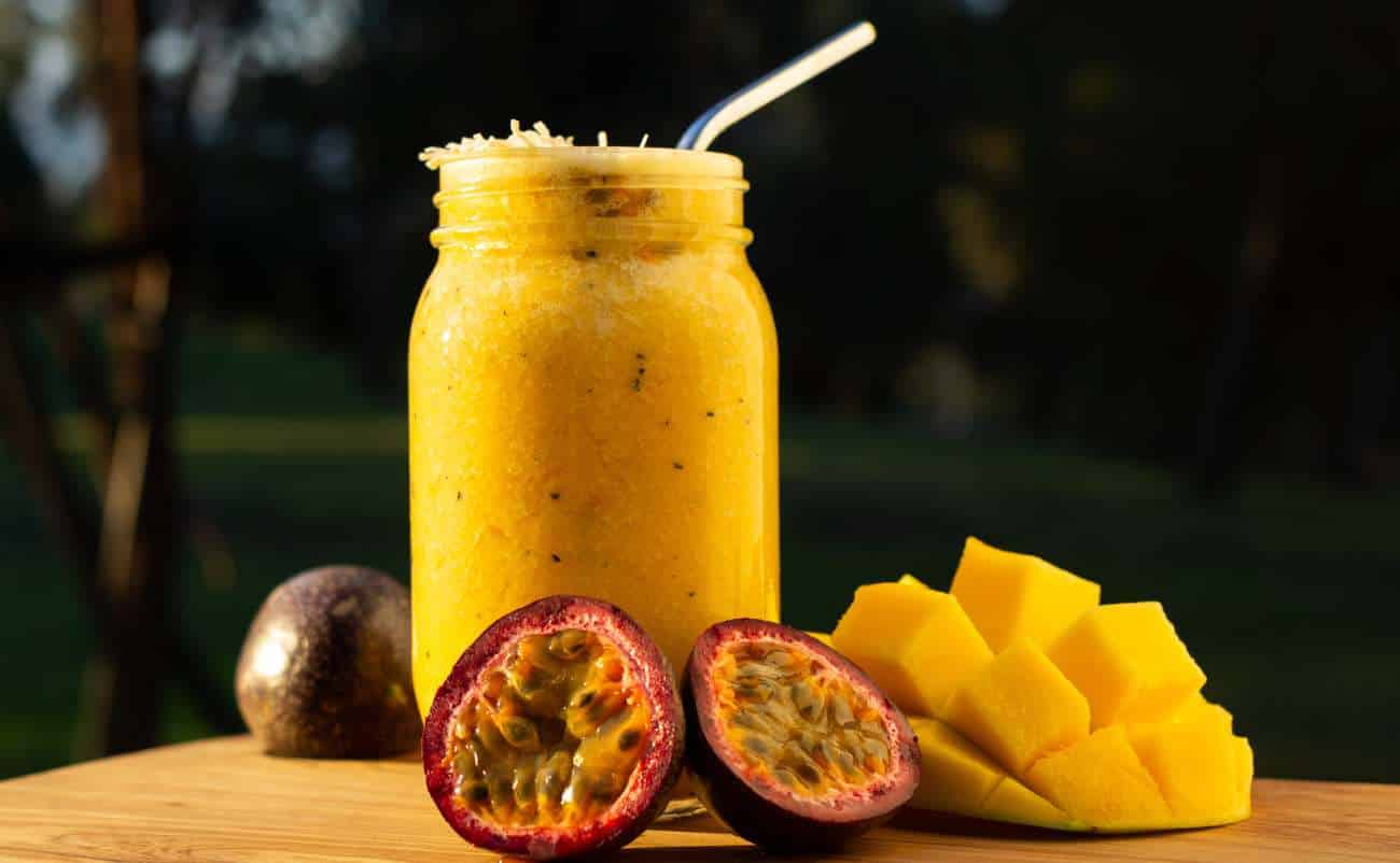 Fresh passion fruit and mango smoothie in a glass jar with a straw, displayed outdoors on a wooden surface.