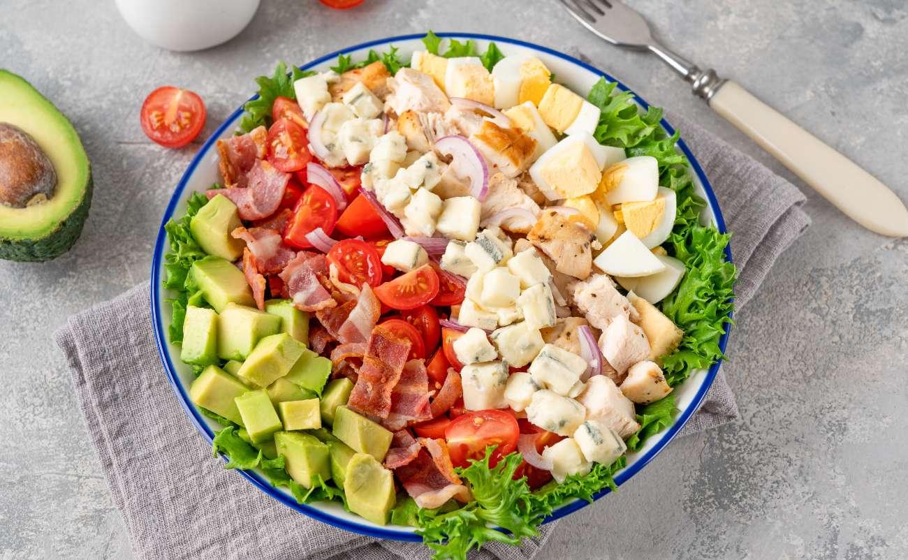 Delicious fresh salad with lettuce, cherry tomatoes, avocado, bacon, boiled eggs, and grilled chicken, garnished with cheese and red onion, served on a gray textured surface.