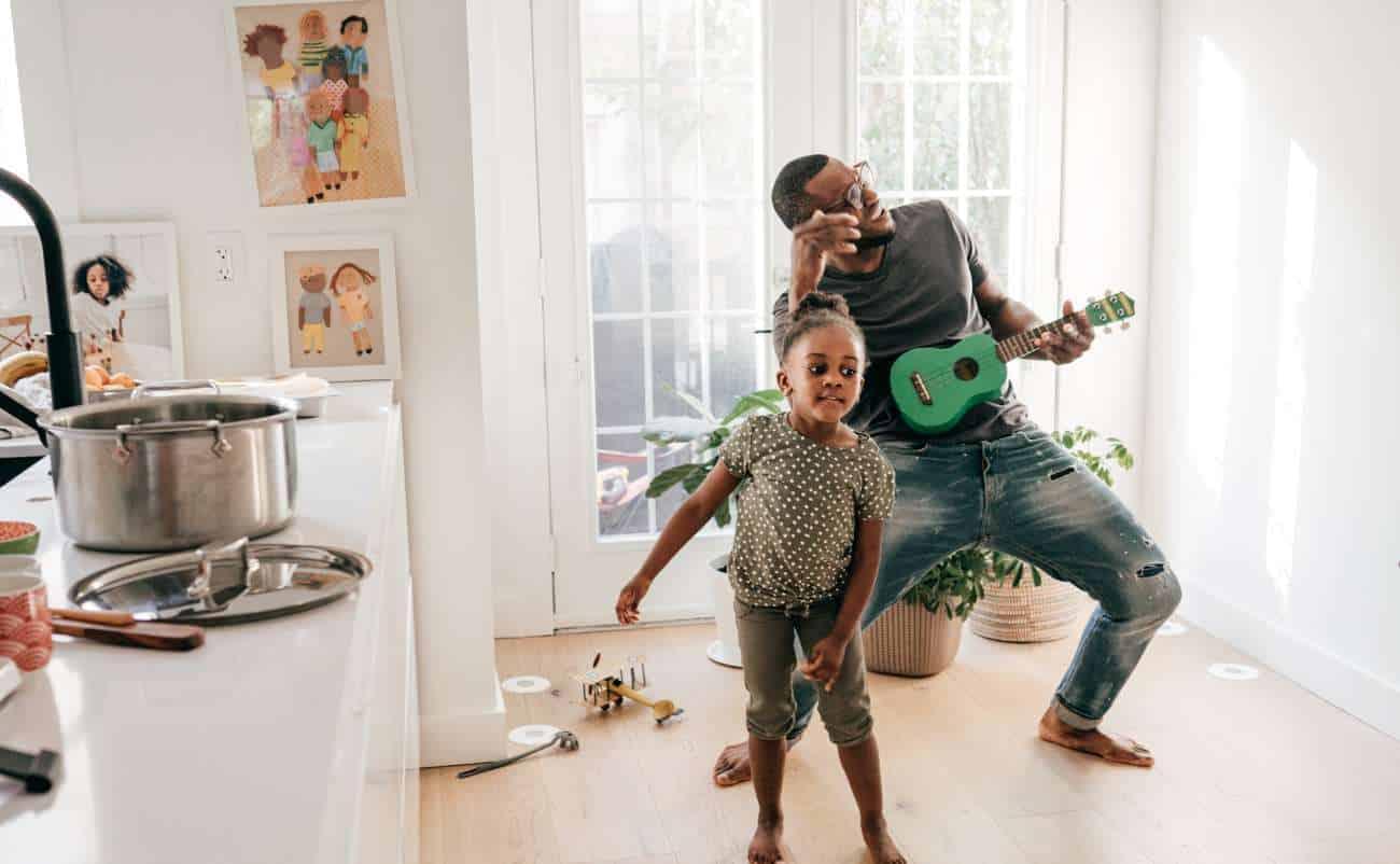 Father playing guitar and singing with daughter dancing in a bright home, creating a joyful and healthy family activity.