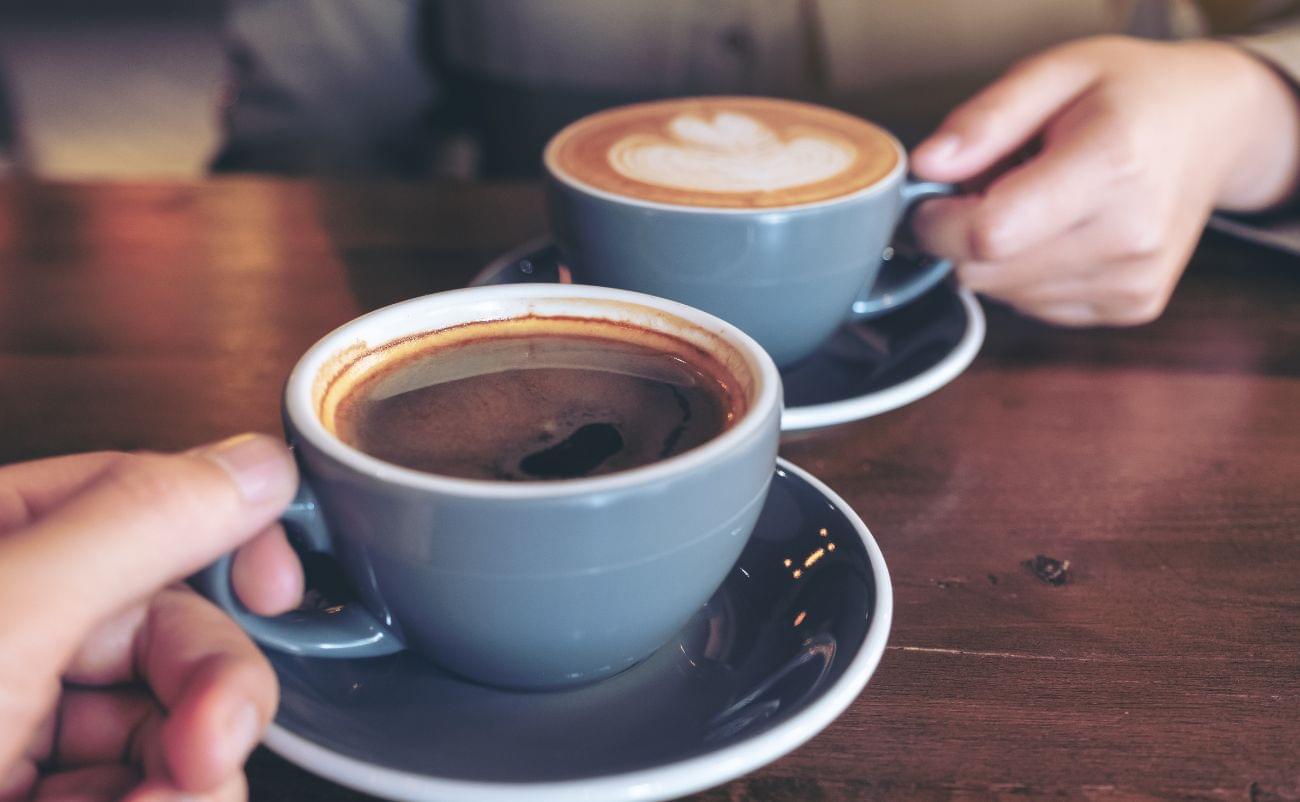 Rich, steaming cups of coffee served in gray ceramic cups on a wooden table, with one person holding a cup and another reaching for their coffee, suggesting a relaxing coffee break or social moment.