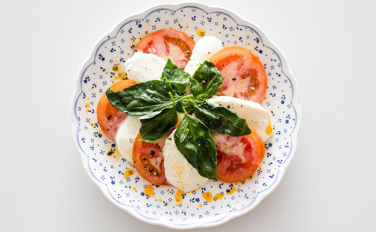 Fresh Caprese salad with ripe tomatoes, mozzarella, basil leaves, olive oil, and black pepper on a decorative white plate.