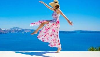 Sun-kissed woman in a flowing floral dress and straw hat enjoying a carefree moment by the sea with scenic blue ocean and sky backdrop.