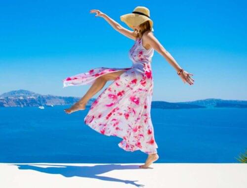 Sun-kissed woman in a flowing floral dress and straw hat enjoying a carefree moment by the sea with scenic blue ocean and sky backdrop.
