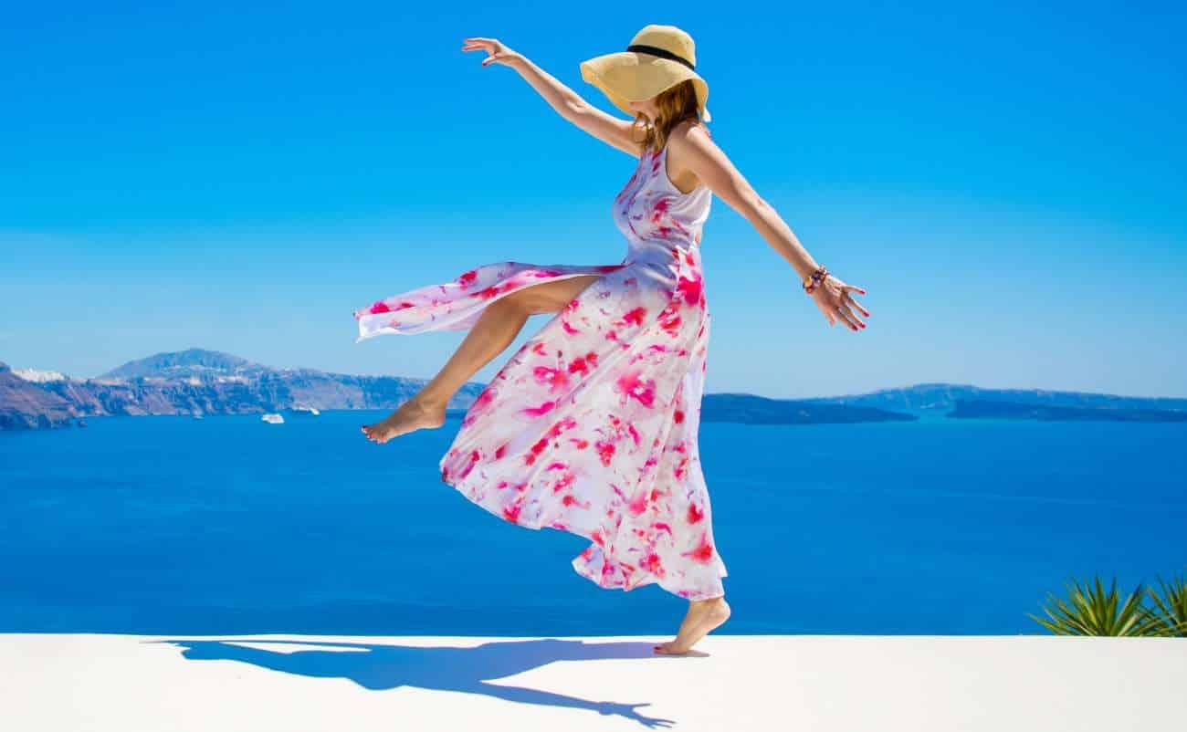 Sun-kissed woman in a flowing floral dress and straw hat enjoying a carefree moment by the sea with scenic blue ocean and sky backdrop.