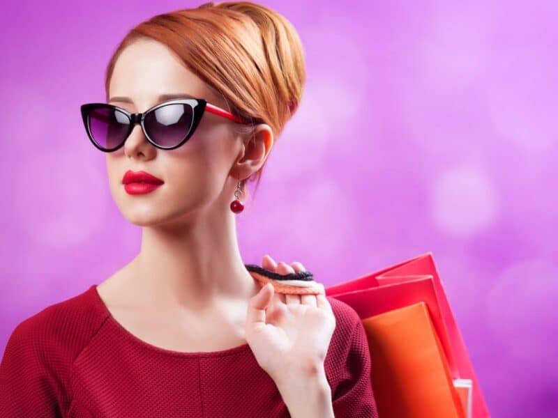 Sassy woman with red hair wearing sunglasses and red lipstick, carrying shopping bags against a vibrant purple background.