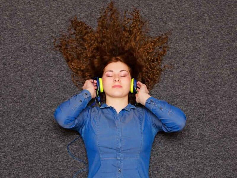 Young woman with curly hair lying on carpet, listening to music with headphones, relaxed and enjoying her workout time at home or gym, promoting stress relief and fitness.