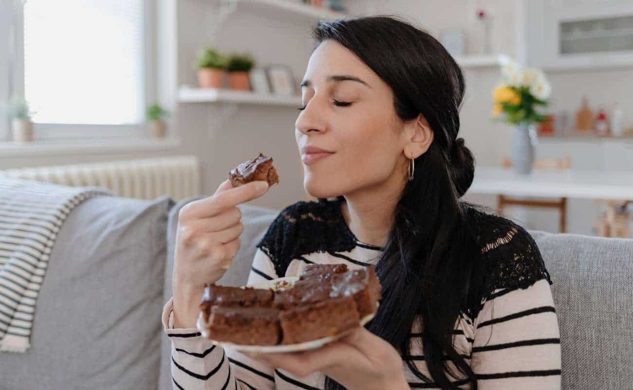 Sweet woman enjoying a bite of chocolate cake while sitting on a sofa in a bright, cozy living room.