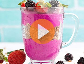 Vibrant pink smoothie in a glass mug topped with fresh berries, garnished with white chocolate shavings over a light turquoise background.