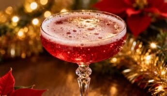 Alt text: A sparkling red holiday cocktail in an elegant glass with a frothy top, surrounded by Christmas decorations like poinsettias and twinkling lights, perfect for festive celebrations.
