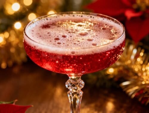 Alt text: A sparkling red holiday cocktail in an elegant glass with a frothy top, surrounded by Christmas decorations like poinsettias and twinkling lights, perfect for festive celebrations.