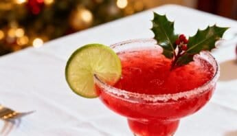 A vibrant red holiday cocktail garnished with a lime slice and holly leaves, served in a salted rim glass on a white tablecloth, perfect for celebrating the season.