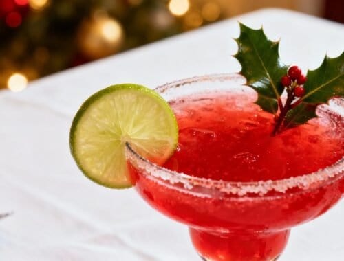 A vibrant red holiday cocktail garnished with a lime slice and holly leaves, served in a salted rim glass on a white tablecloth, perfect for celebrating the season.