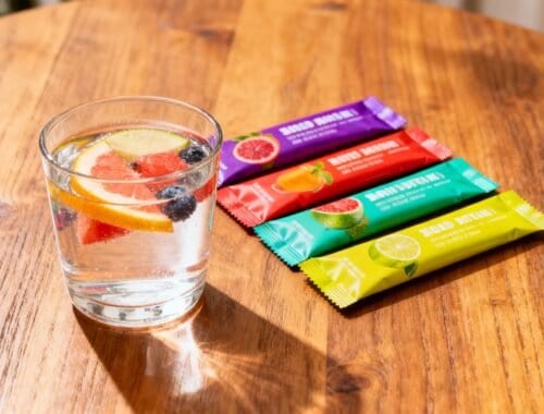Refreshing glass of infused water with lemon, berries, and grapefruit on a wooden table, with colorful healthy snack bars nearby, promoting hydration and nutritious snacking options.