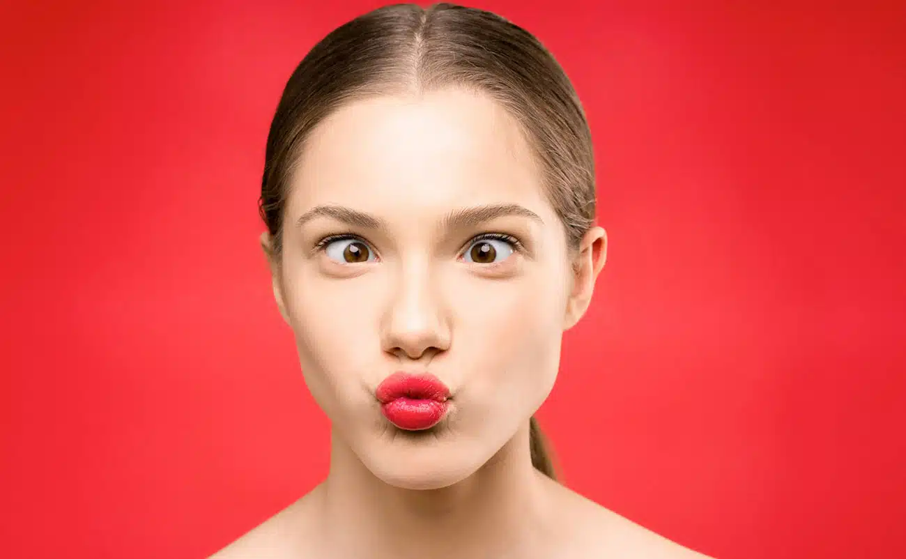 Smiling young woman with puckered lips making a playful kiss face against a vibrant red background.
