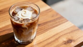 Creamy iced coffee with swirls of milk and whipped cream in a clear glass on a wooden table.