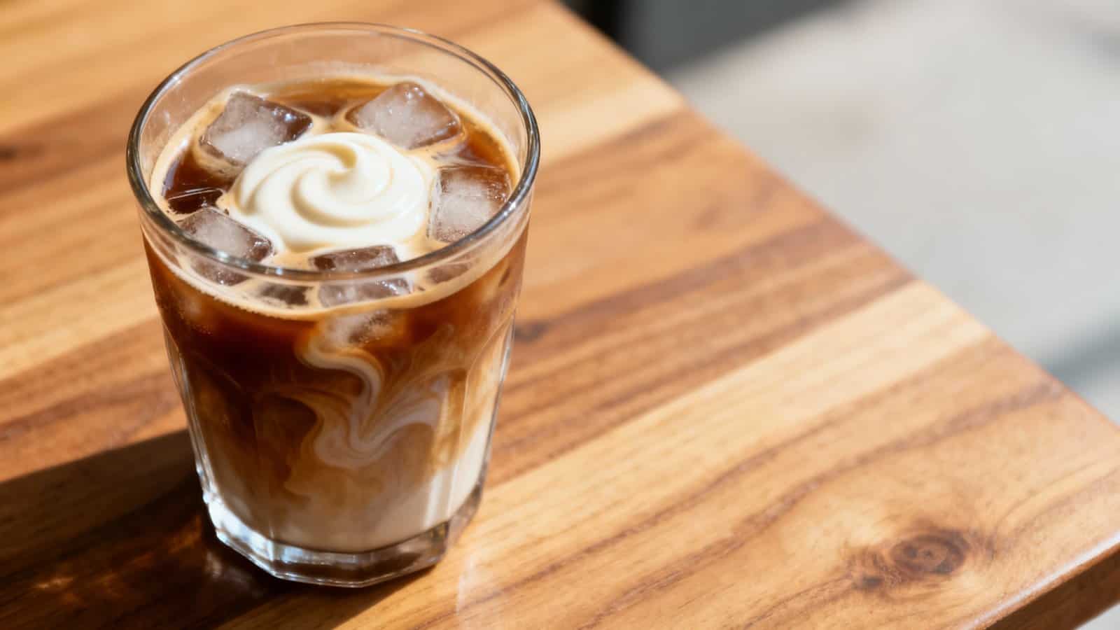 Creamy iced coffee with swirls of milk and whipped cream in a clear glass on a wooden table.