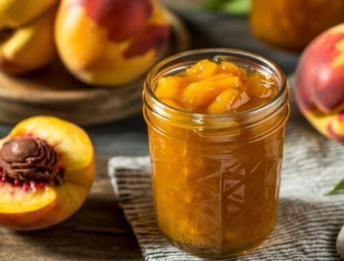 Fresh peach preserves in a jar surrounded by ripe, colorful peaches on a rustic wooden table. Perfect for health-conscious eating and homemade fruit spreads.