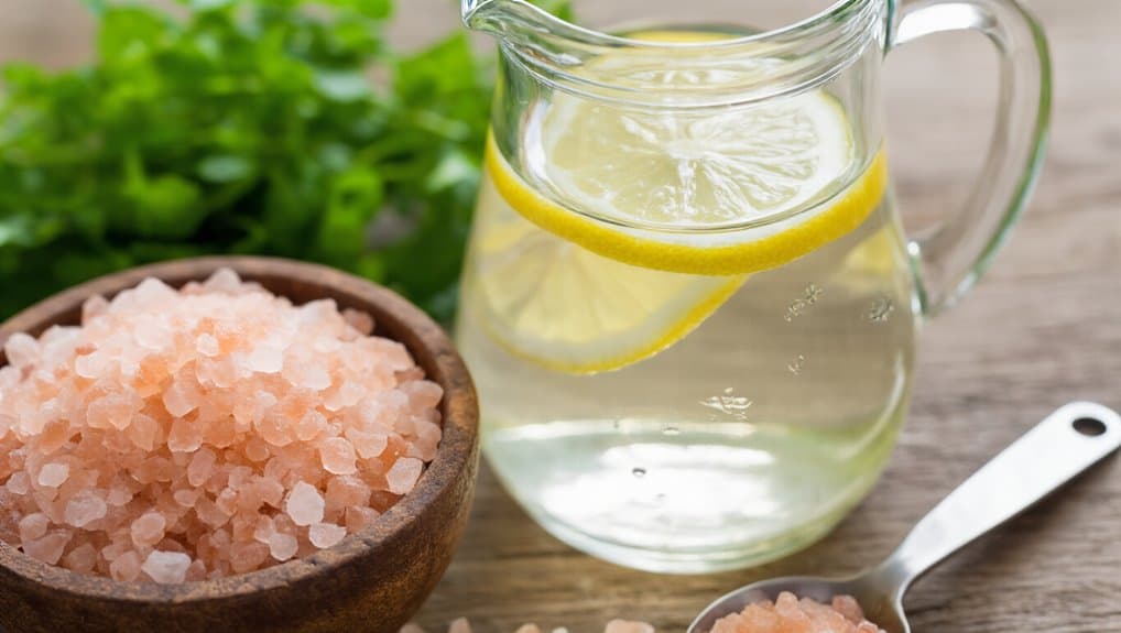 Natural pink Himalayan salt in a wooden bowl, a glass pitcher of infused lemon water, and fresh green herbs on a rustic wooden surface promote healthy hydration and mineral intake.