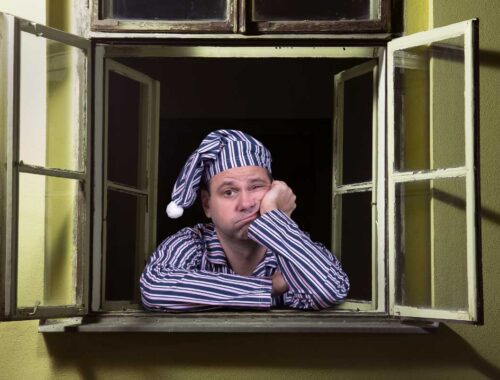 Restless man in pajamas and nightcap sitting at an open window, looking tired and bored, with a disappointed expression, suggesting sleep deprivation or insomnia.