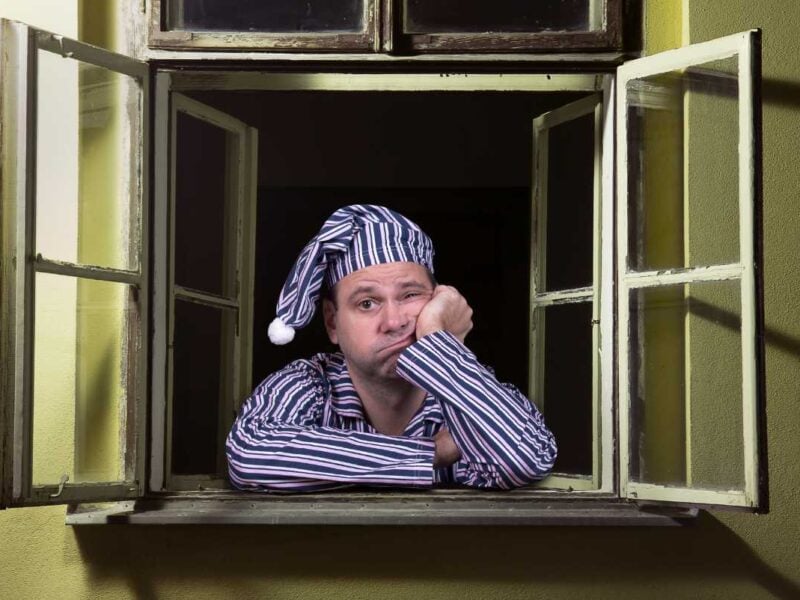 Restless man in pajamas and nightcap sitting at an open window, looking tired and bored, with a disappointed expression, suggesting sleep deprivation or insomnia.