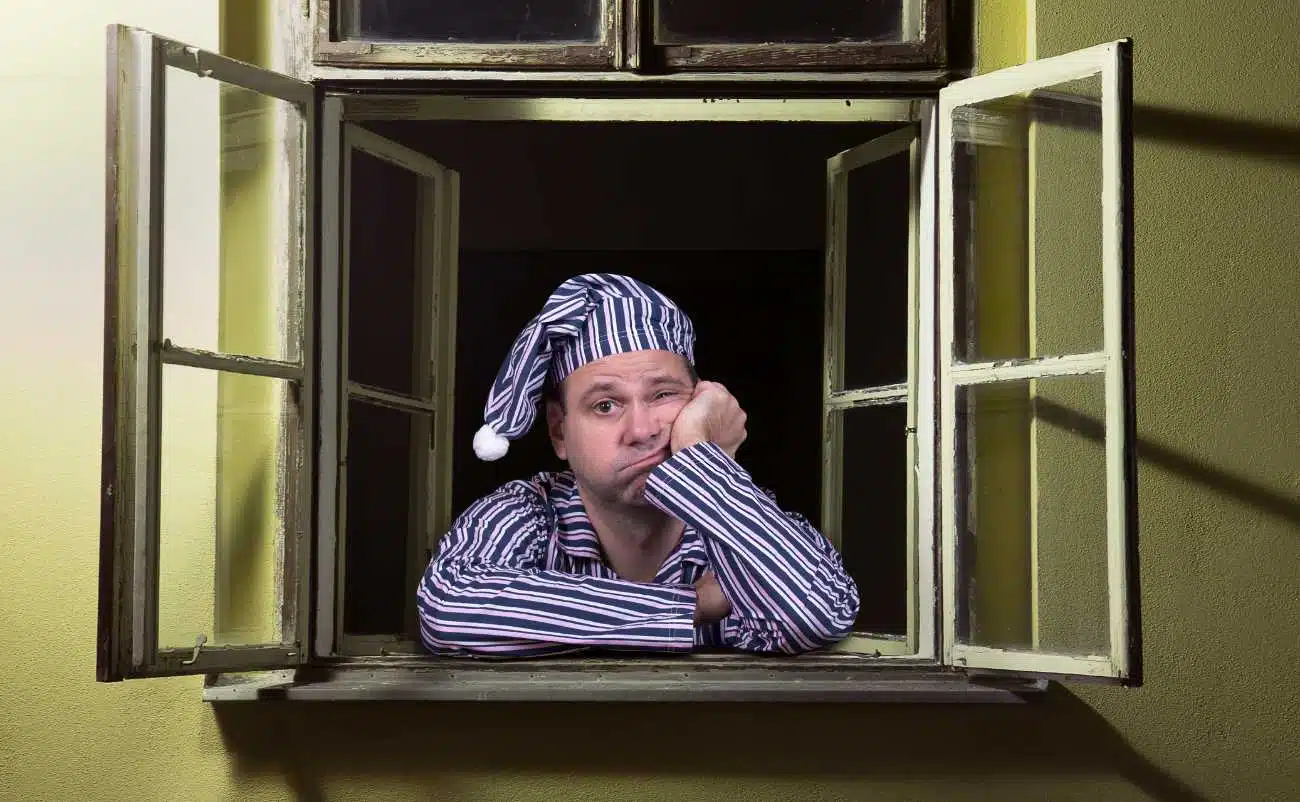 Restless man in pajamas and nightcap sitting at an open window, looking tired and bored, with a disappointed expression, suggesting sleep deprivation or insomnia.