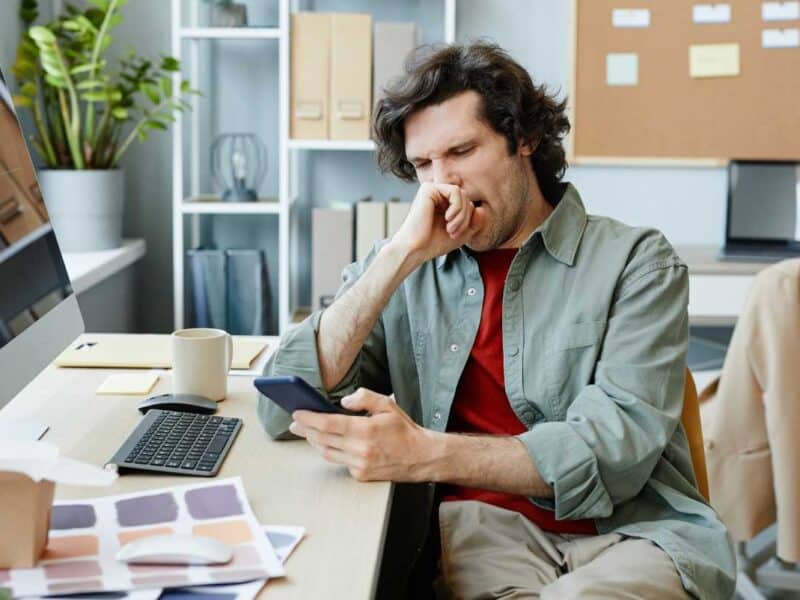 Feeling unwell man coughing at office desk looking at smartphone health and wellness concerns healthcare stress support concept.