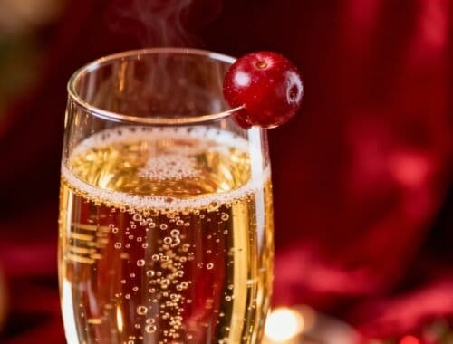 A glass of sparkling champagne garnished with a fresh red berry, perfect for holiday celebrations and special occasions. The bubbly drink is set against a festive, blurred background, evoking a joyful, elegant atmosphere.
