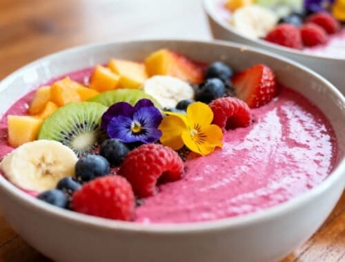 Fresh fruit smoothie bowl topped with colorful berries, kiwi, banana, edible flowers, and diced mango, served in a white bowl on a wooden surface, promoting healthy eating habits.