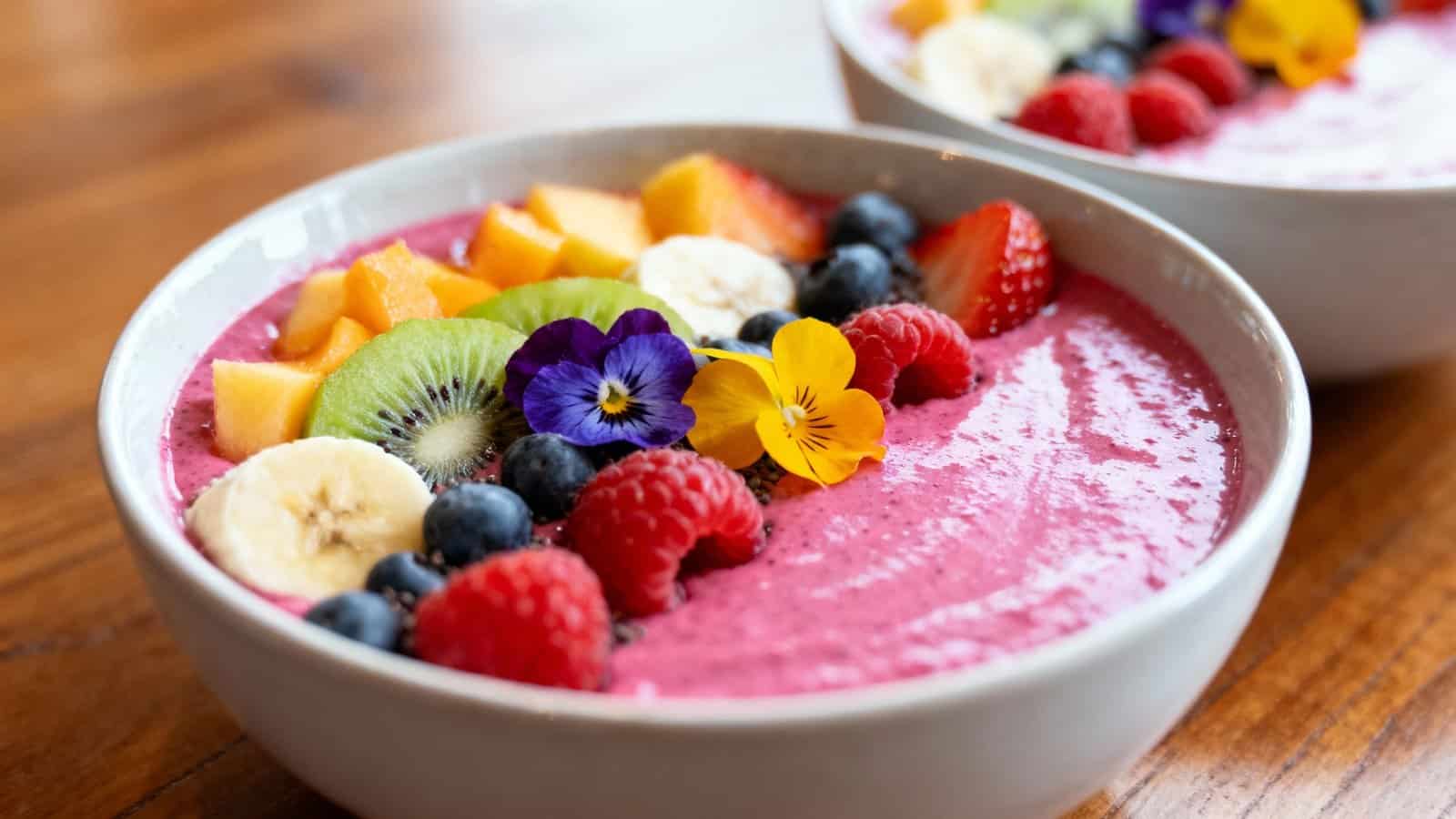 Fresh fruit smoothie bowl topped with colorful berries, kiwi, banana, edible flowers, and diced mango, served in a white bowl on a wooden surface, promoting healthy eating habits.