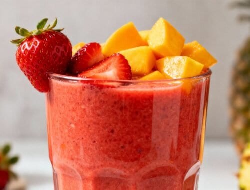 Fresh strawberry and mango smoothie in a clear glass, topped with sliced strawberries and mango chunks, with pineapple and strawberries in the background.