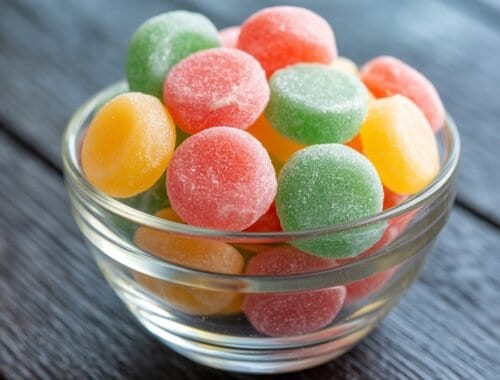 Colorful fruit gummies in a glass bowl on a wooden surface, showcasing healthy snack options for fitness enthusiasts.