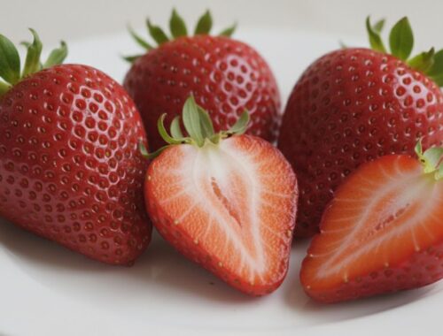 Close-up of ripe strawberries, some halved to show their vibrant red interior and seeds, placed on a white plate. The strawberries are fresh, ripe, and perfect for healthy snacks or desserts.