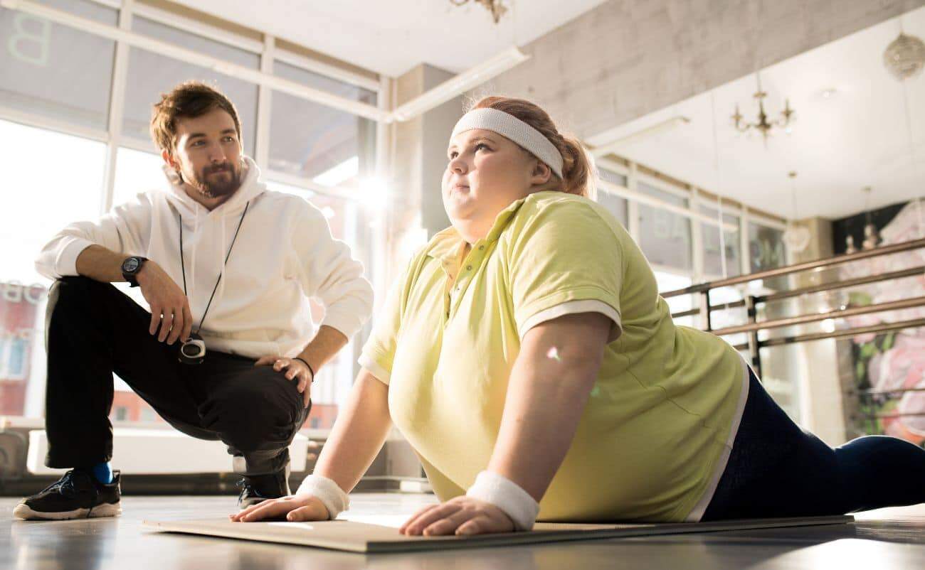 "Female overweight client performing a plank exercise guided by her fitness trainer in a bright gym.".