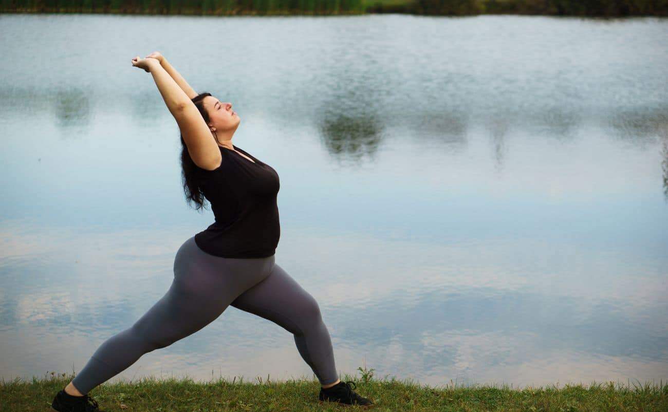 Yoga practice for weight loss by a woman doing a Warrior pose outdoors near a lake.