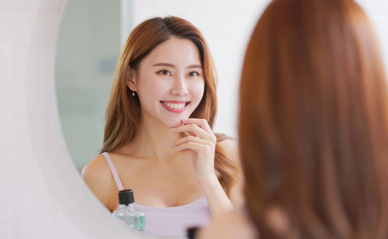 Smooth young woman with red hair looking at herself in the mirror, smiling, in a bright bathroom setting.