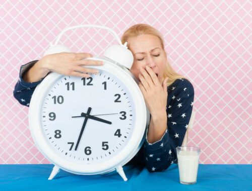 Too much sleep inducing fatigue, woman yawning next to a large alarm clock and glass of milk on blue table, pink patterned background.
