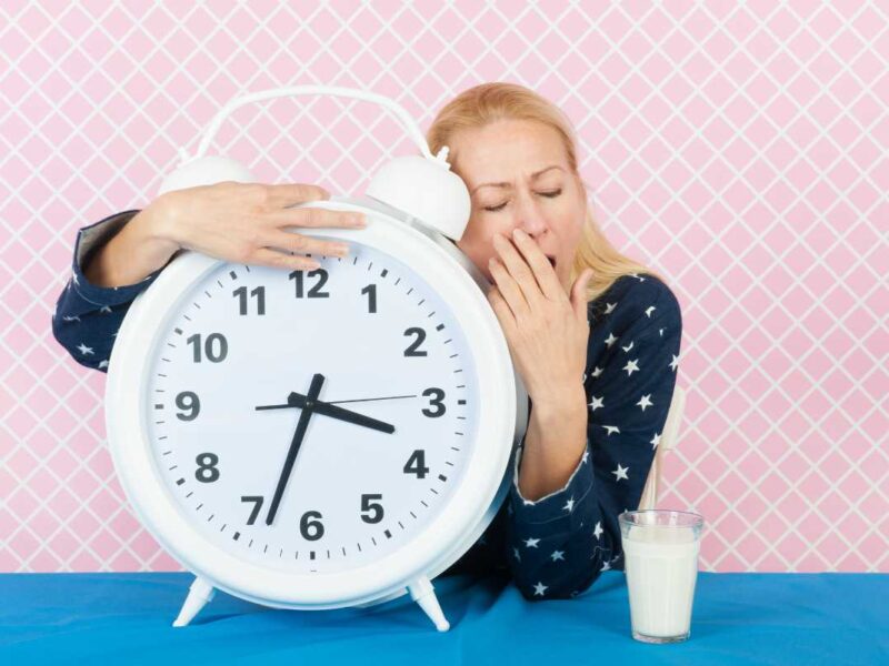 Too much sleep inducing fatigue, woman yawning next to a large alarm clock and glass of milk on blue table, pink patterned background.