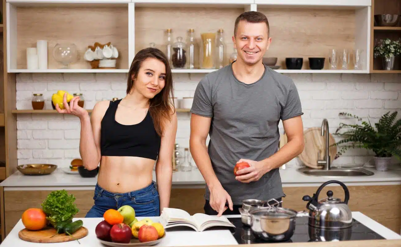 Fresh healthy fruits and vegetables on kitchen counter with a smiling man and woman preparing a meal together.